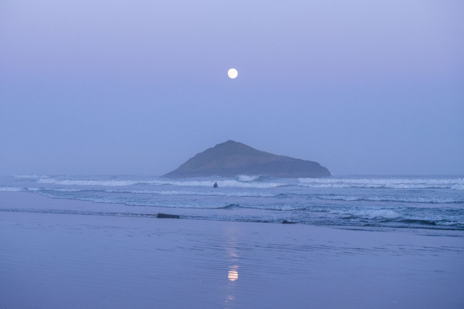 Full moon rising over a misty beach with an island silhouette reflecting on calm waters at dusk.