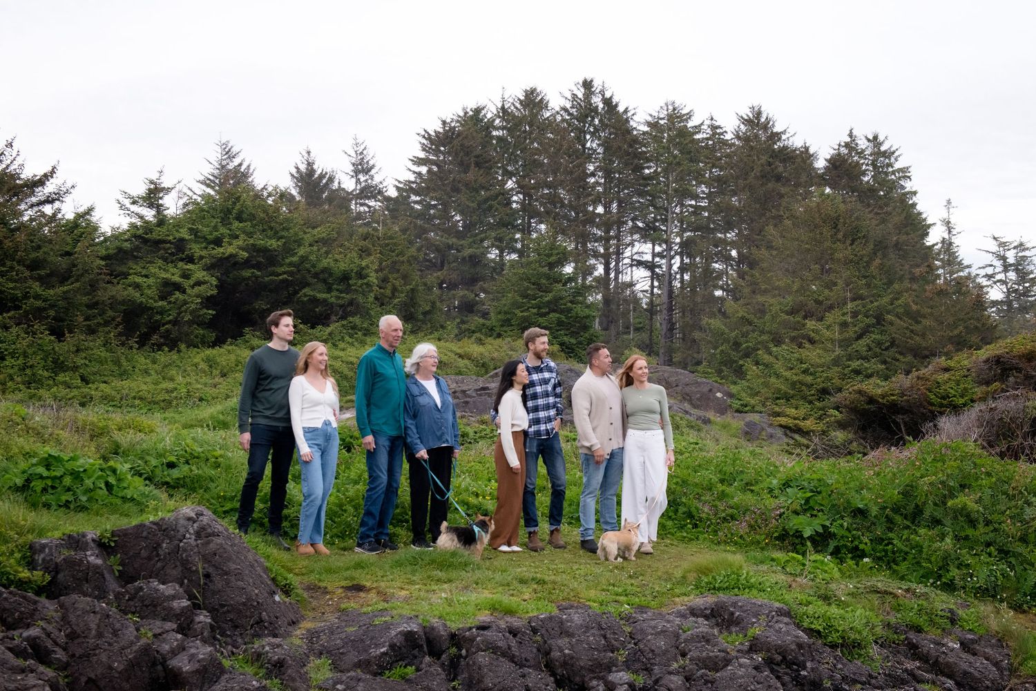 People gathered on a grassy oceanside bluff with evergreen forest and cloudy skies in the distance.