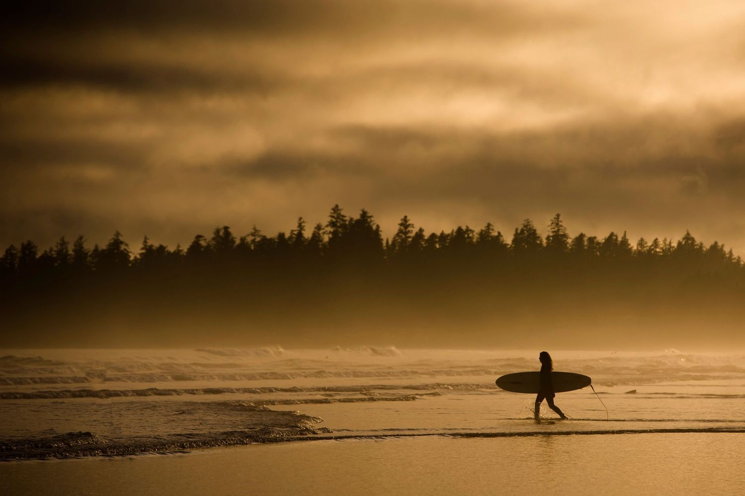 Silhouette of surfer walking along beach at sunset with dramatic orange sky and ocean waves.