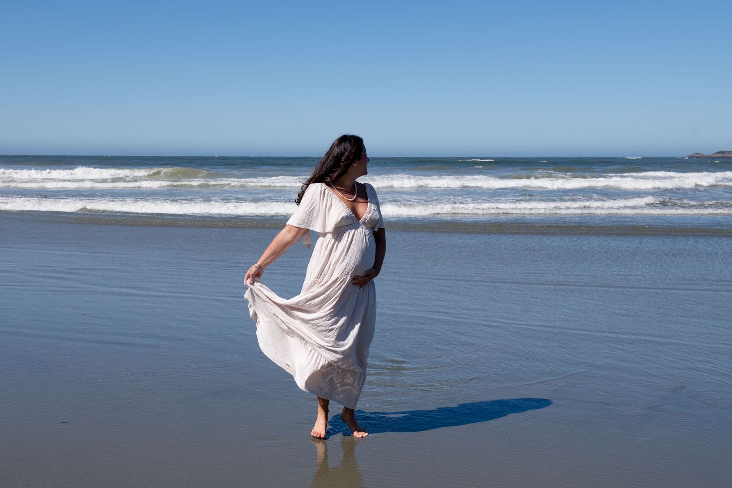 Flowing white dress catches the ocean breeze during beach maternity photos.