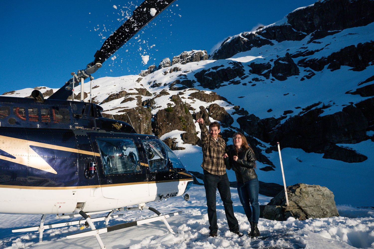 Couple posing together in front of a helicopter on a snowy mountain as rotor blades kick up snow around them.