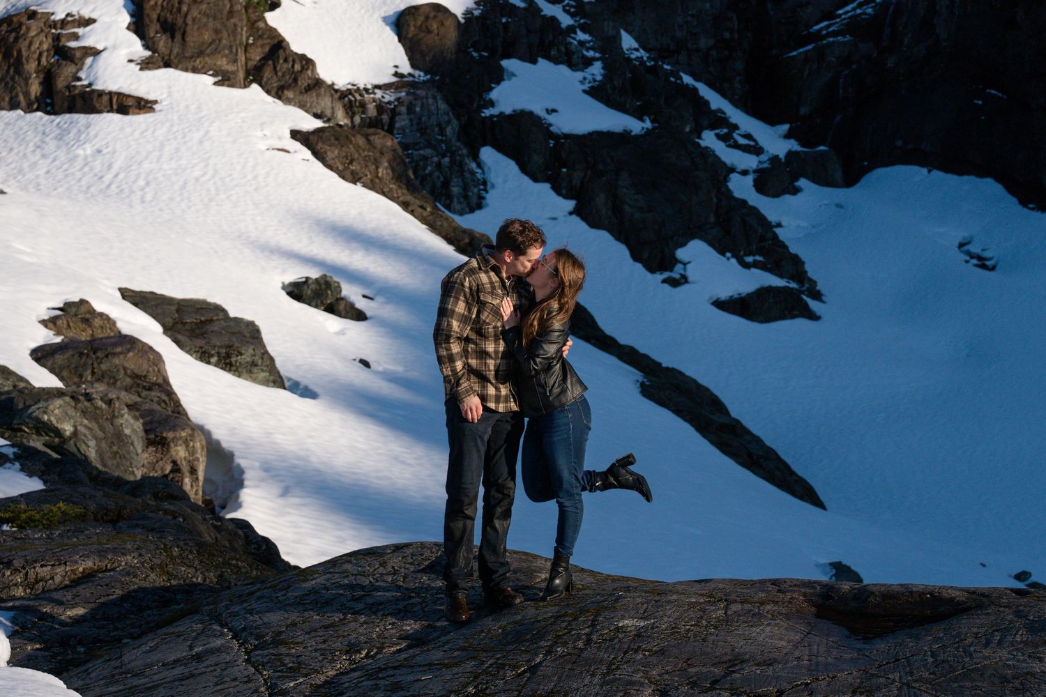 Couple sharing a romantic kiss on snowy mountain rocks during a winter adventure engagement photo session.