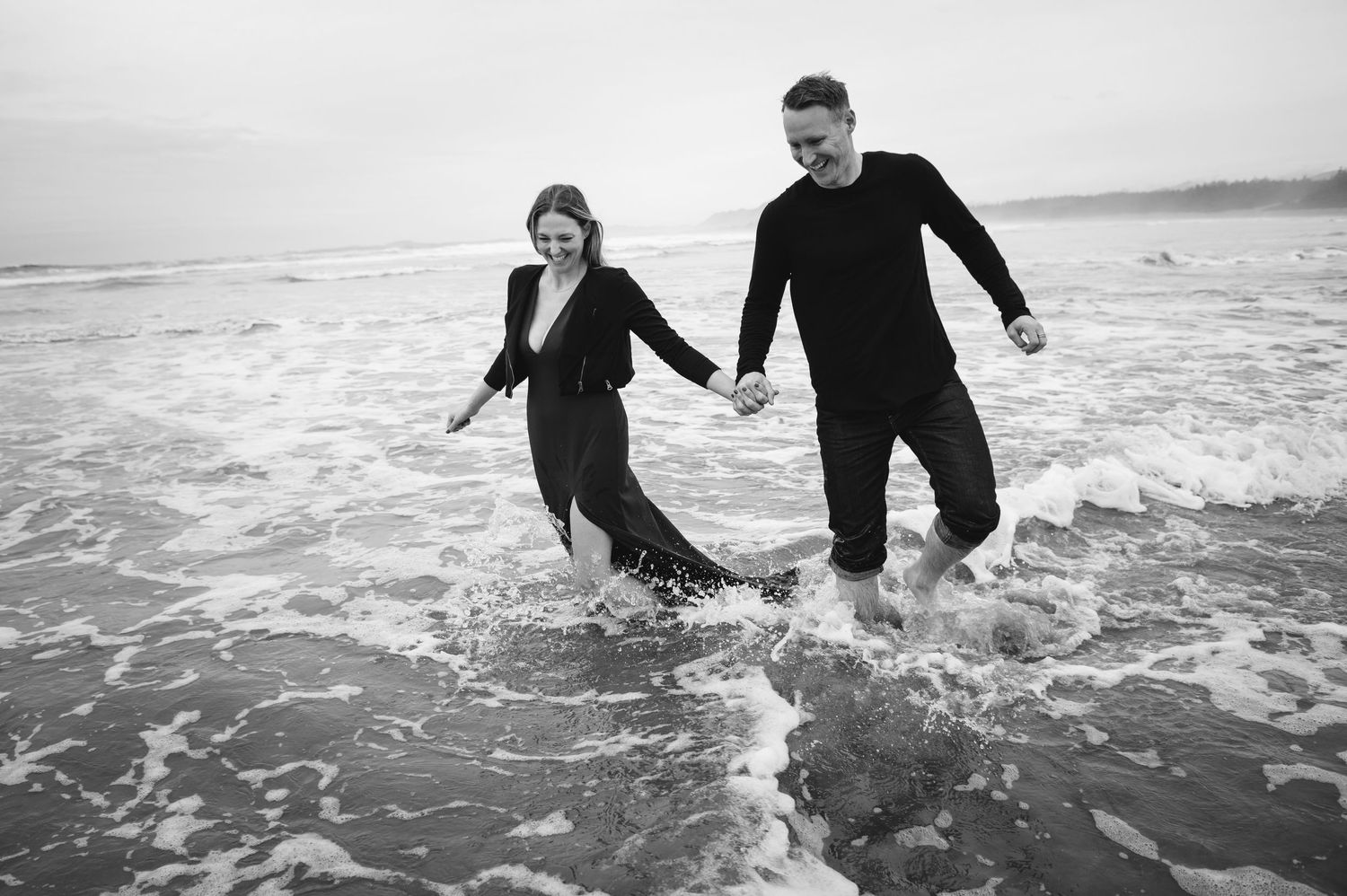 A couple holds hands while splashing through ocean waves in a black and white photo on a moody beach.