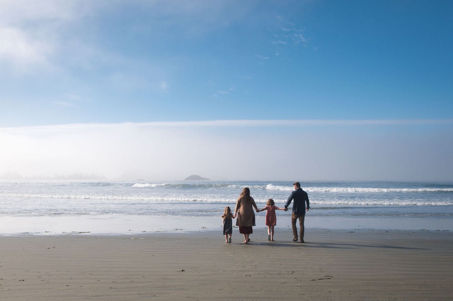 Silhouettes of people walking along a wide sandy beach with mountains in the distance.