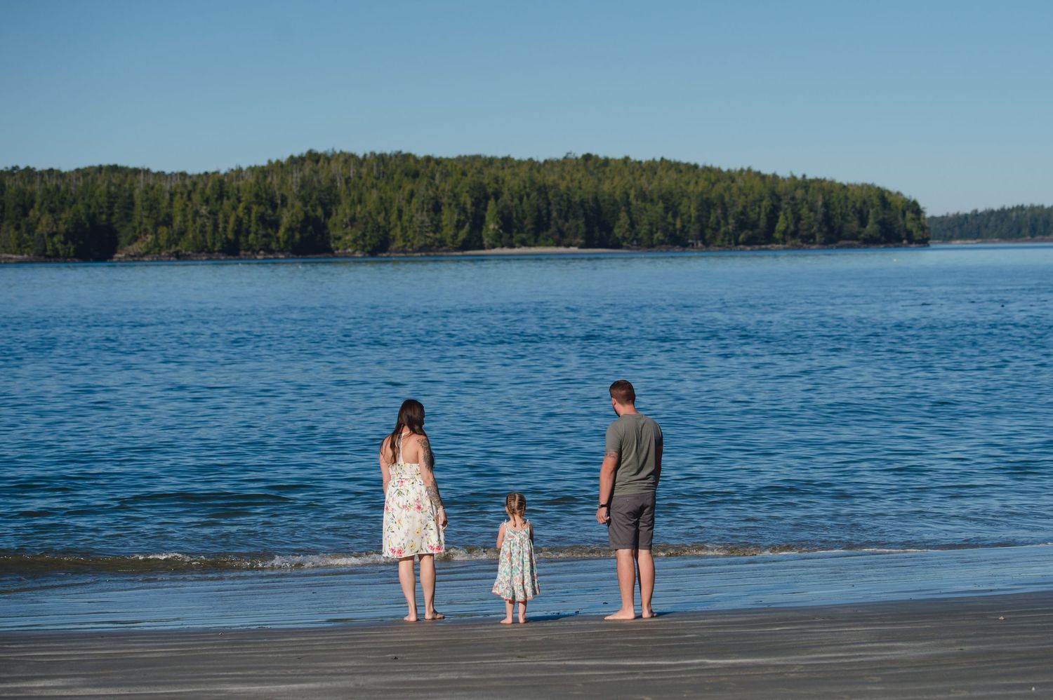 Family walking along scenic beach with evergreen forest and ocean views on sunny day.