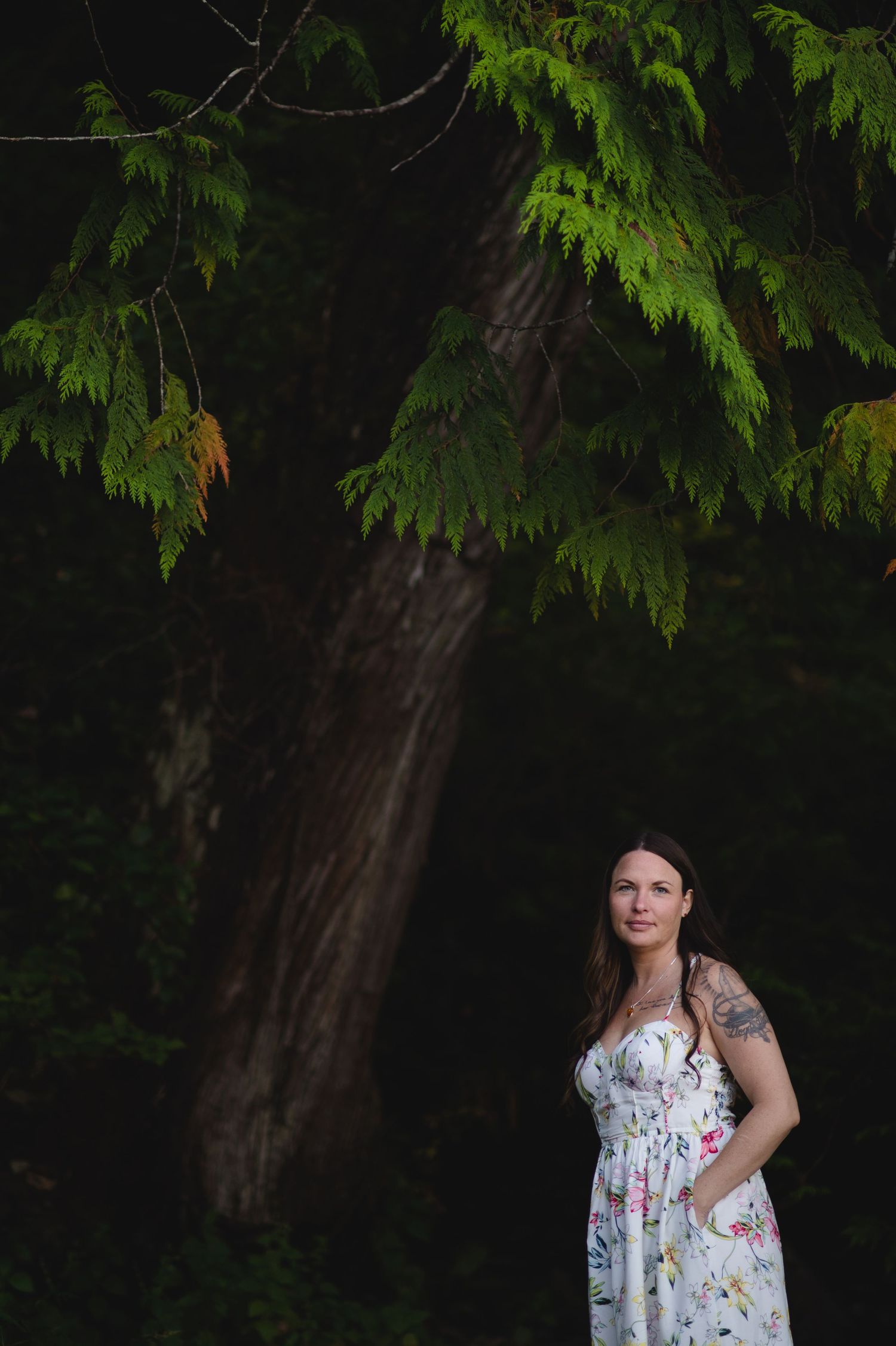 A woman in a white floral dress poses against a backdrop of evergreen trees.