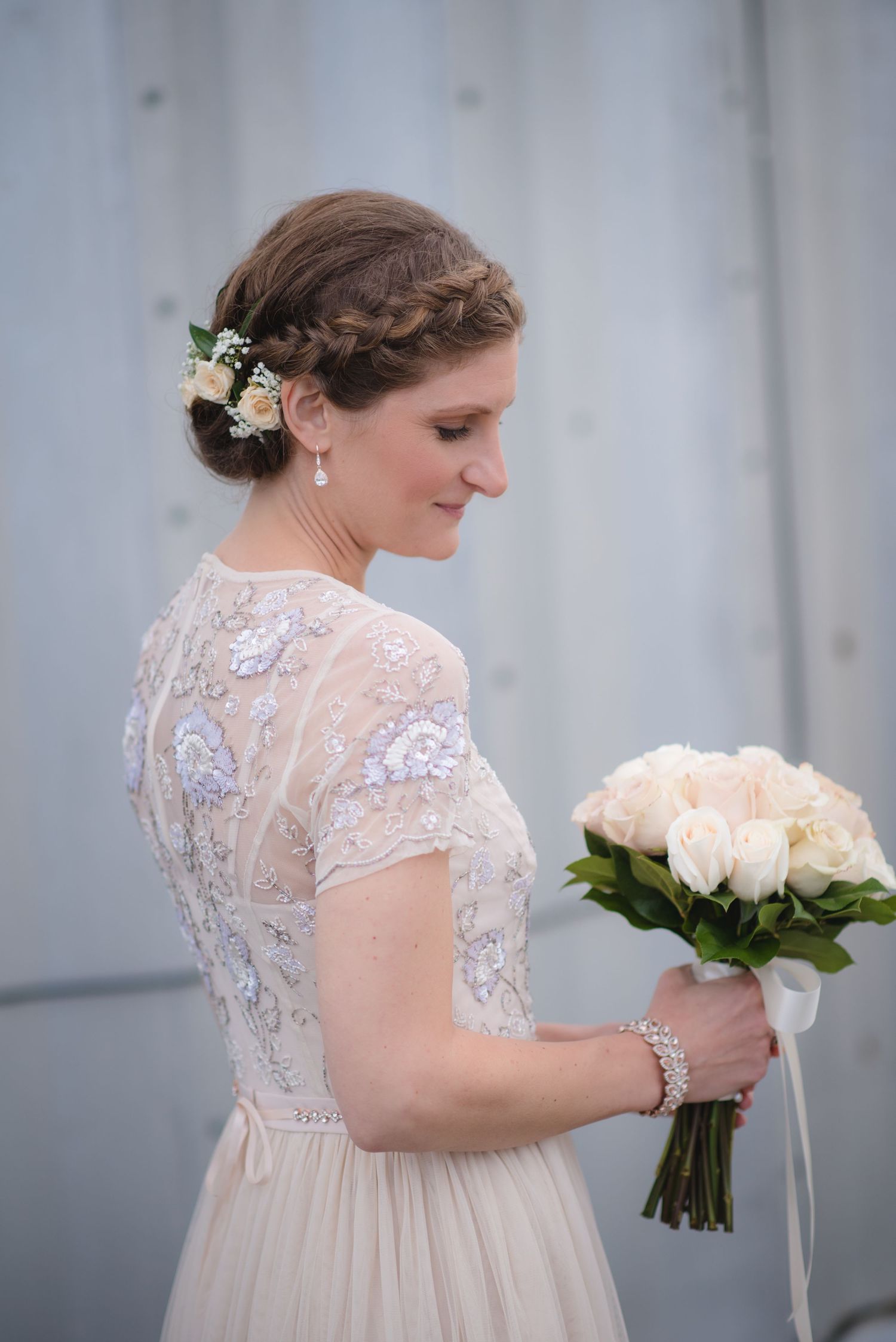 Bride in lace wedding dress holds white rose bouquet against grey backdrop.