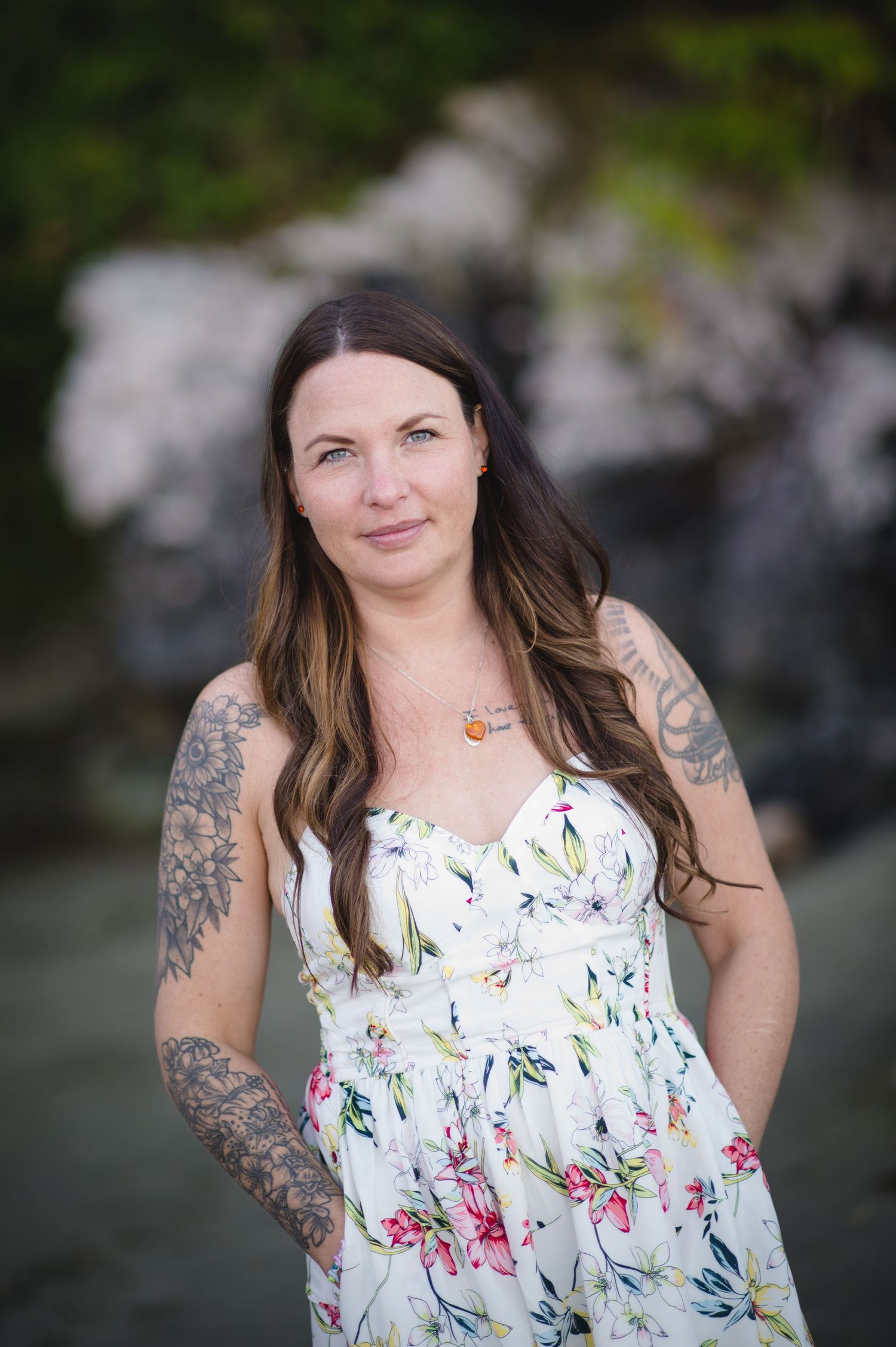 A woman in a white floral dress poses against rocky coastal backdrop with visible tattoos.