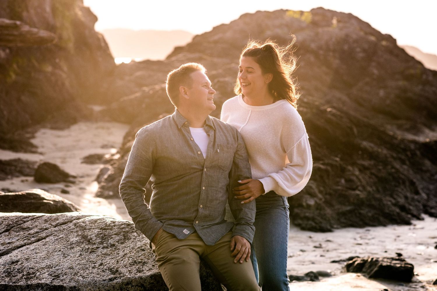 A couple shares a laugh while sitting on dark rocky cliffs by the ocean.