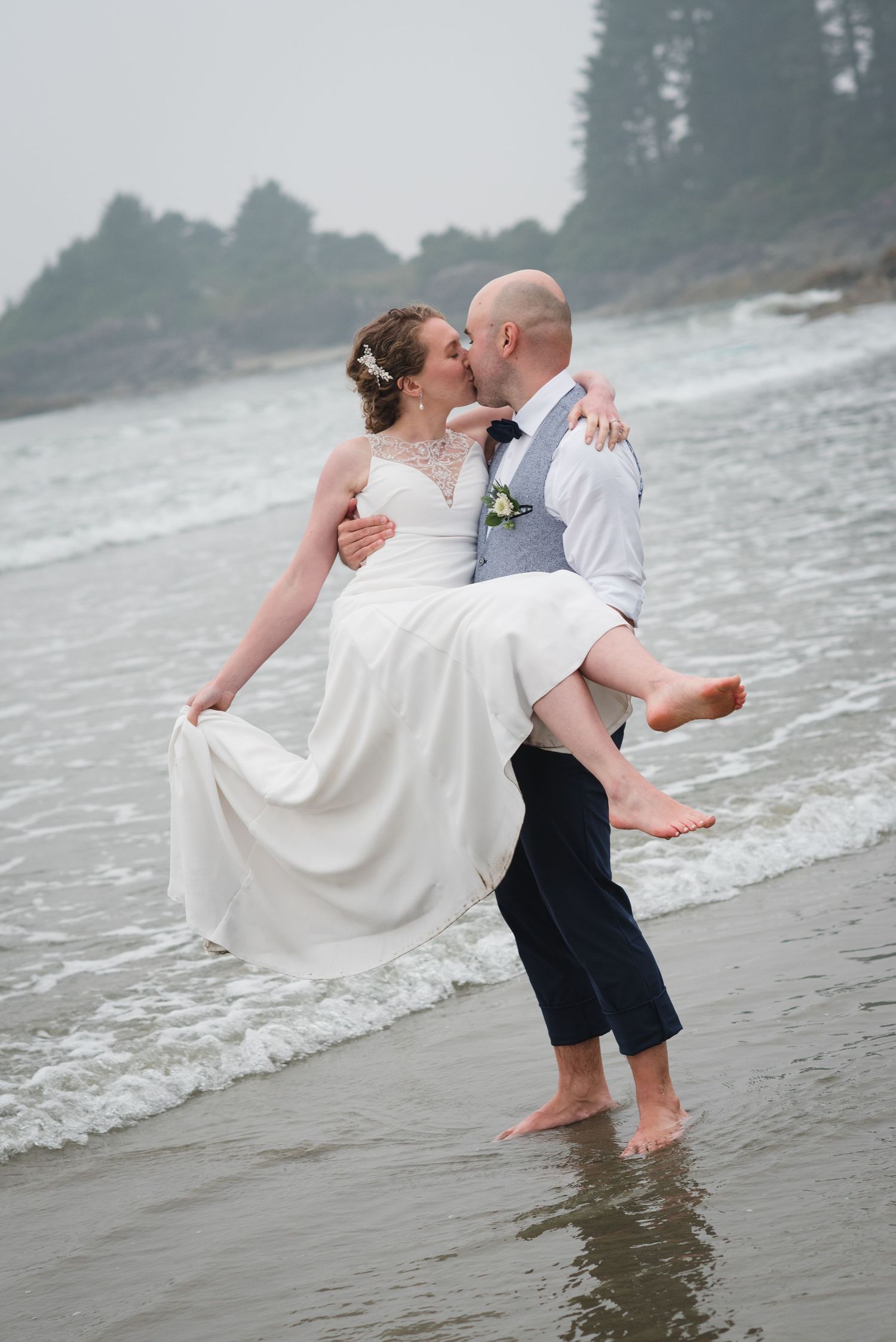 Romantic couple in wedding attire dancing and twirling together on a misty beach shoreline.
