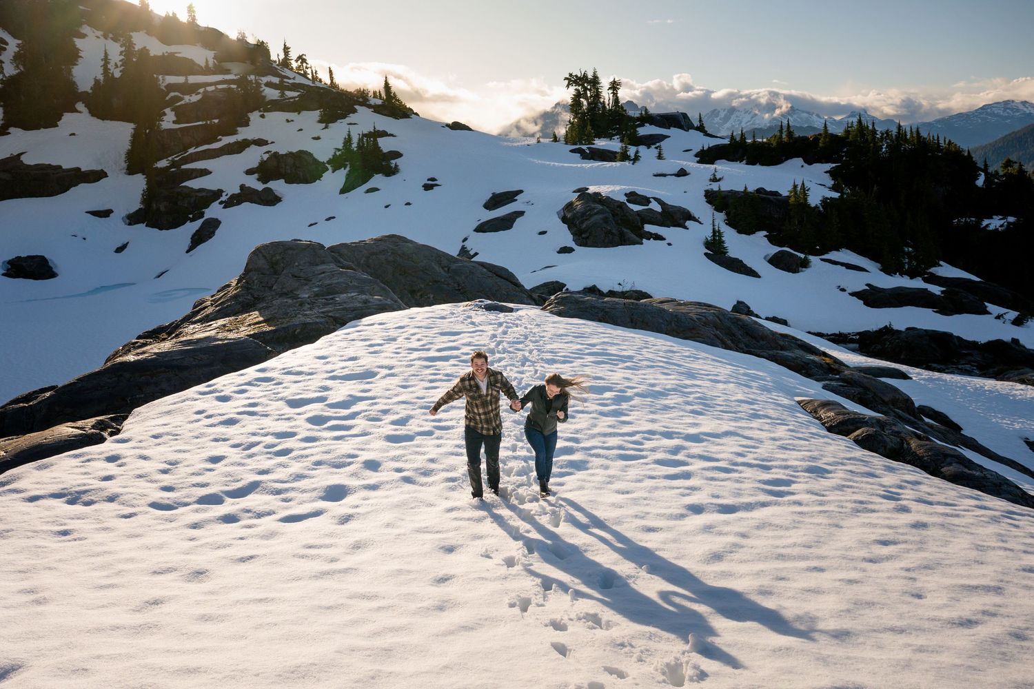Two people holding hands while walking up a snowy mountain slope at sunset with rocky peaks in the background.
