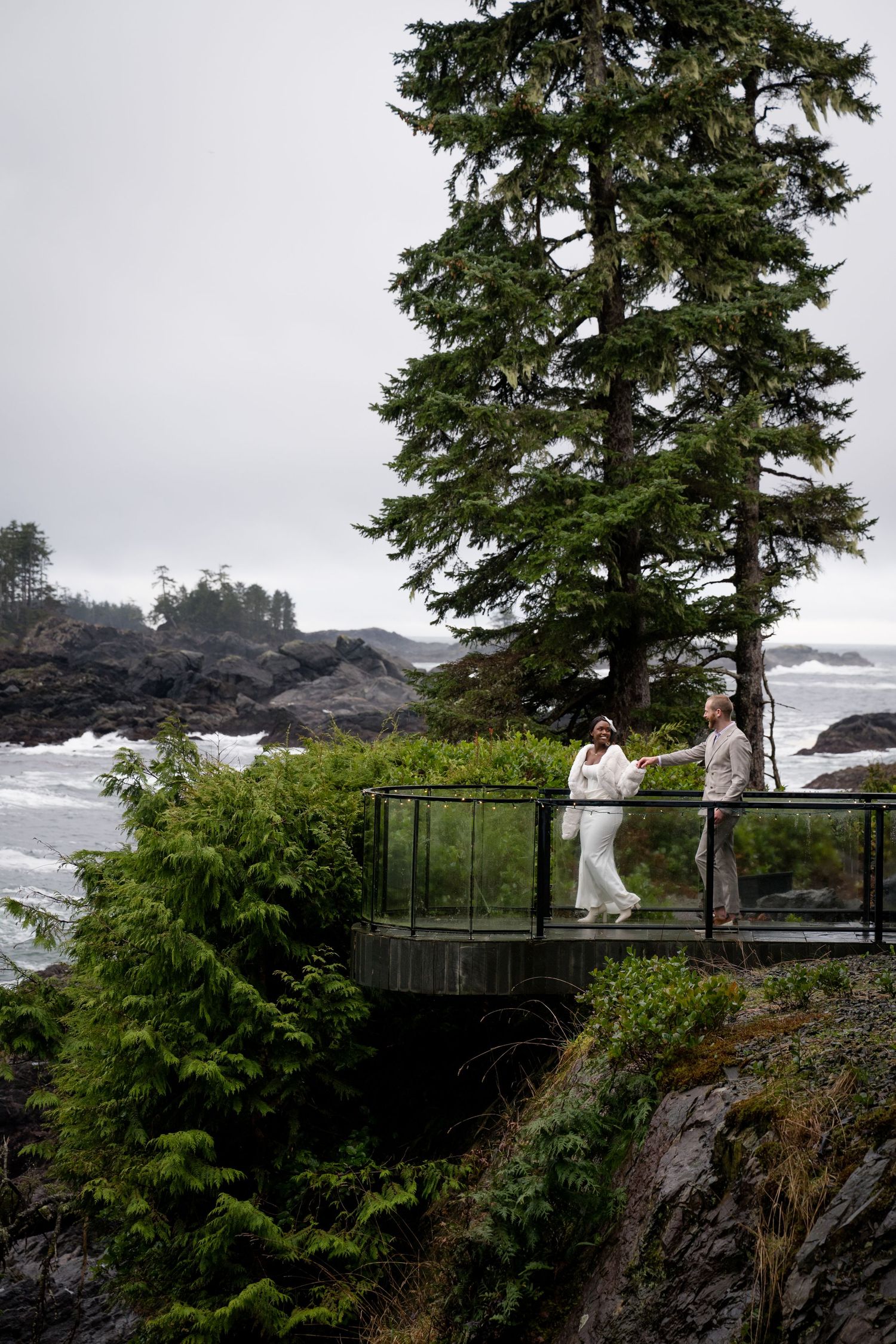A couple stands on a scenic viewing platform overlooking rocky coastal cliffs and evergreen trees in the Pacific Northwest.