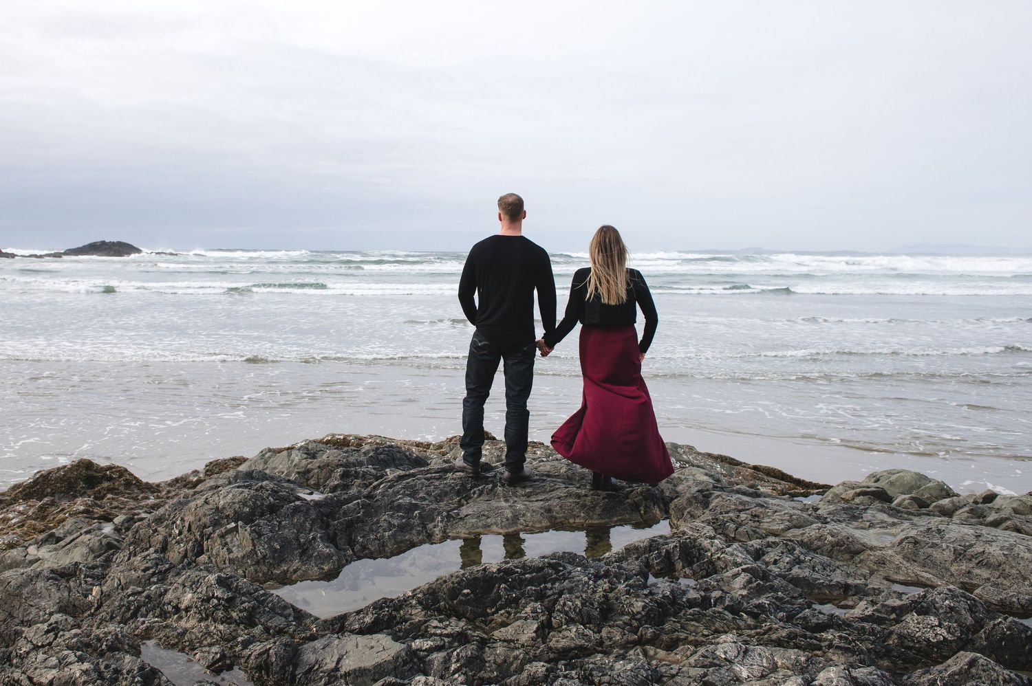 Couple standing on rocky coastal outcrop overlooking dramatic ocean waves.