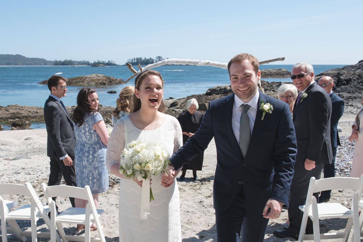 Wedding ceremony in progress on a scenic coastal beach with rocky outcrops and blue water.