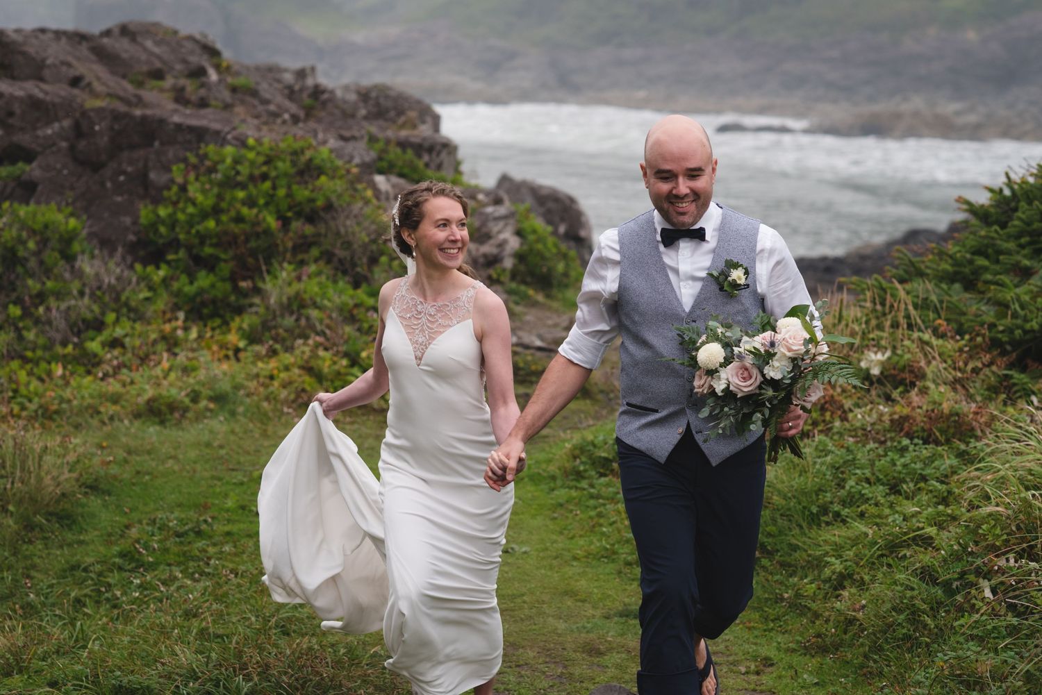 A couple runs playfully along a coastal path with dramatic ocean views in the background.