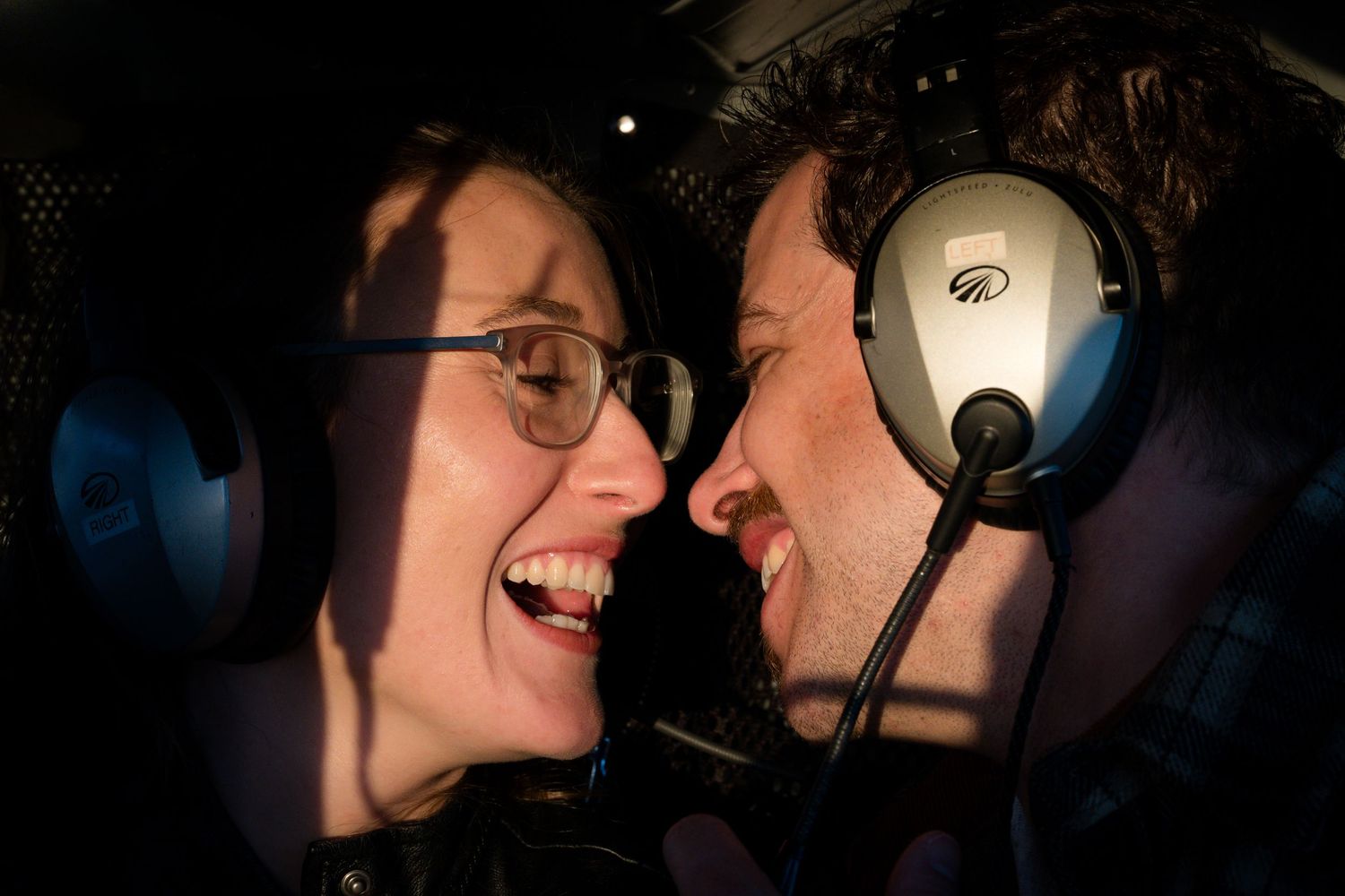 Happy couple laughing together wearing aviation headsets inside a helicopter during romantic mountain flight.