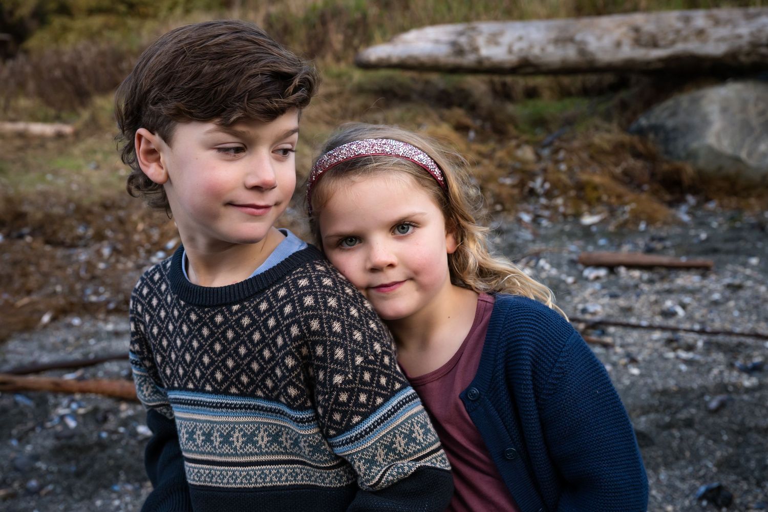 Two children in warm sweaters share a moment together on the rocky shoreline.