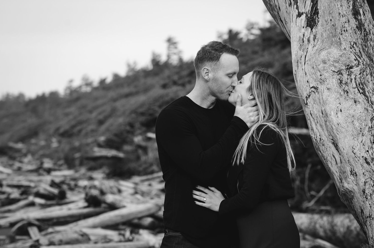 Couple sharing intimate moment near large driftwood on moody beach.
