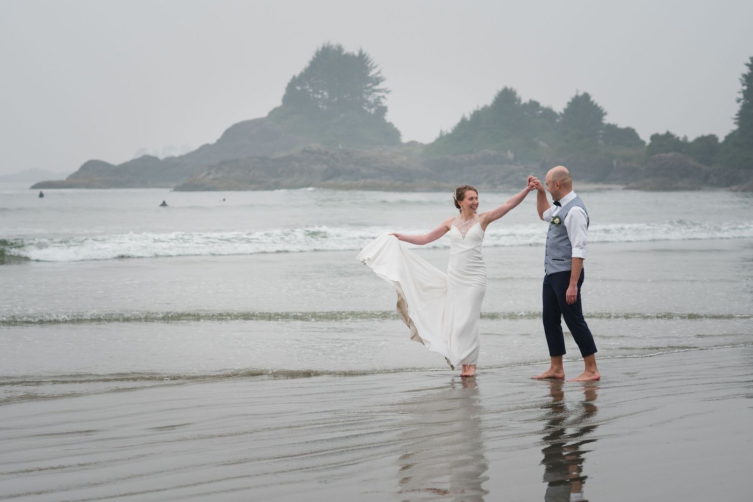 A couple in wedding attire walks along a misty beach shoreline while waves crash behind them.