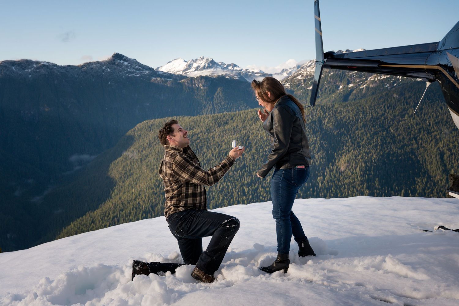 Man proposing to woman on snowy mountain peak with scenic valley and snow-capped mountains in background.