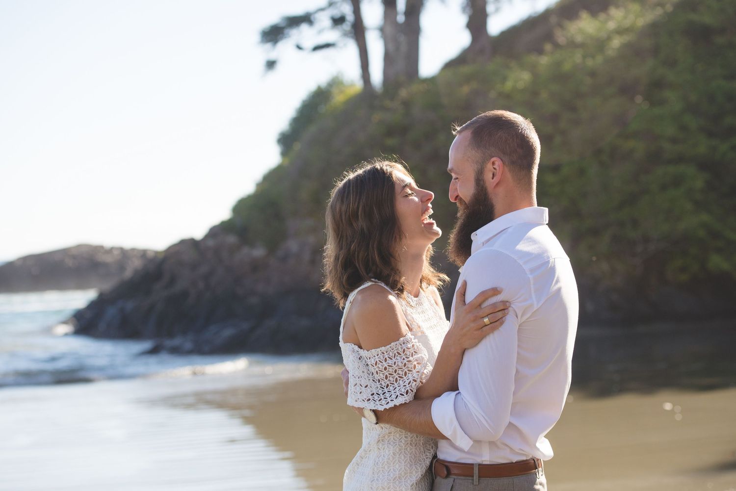 Romantic coastal engagement session with couple dancing on sandy beach near rocky cliffs.