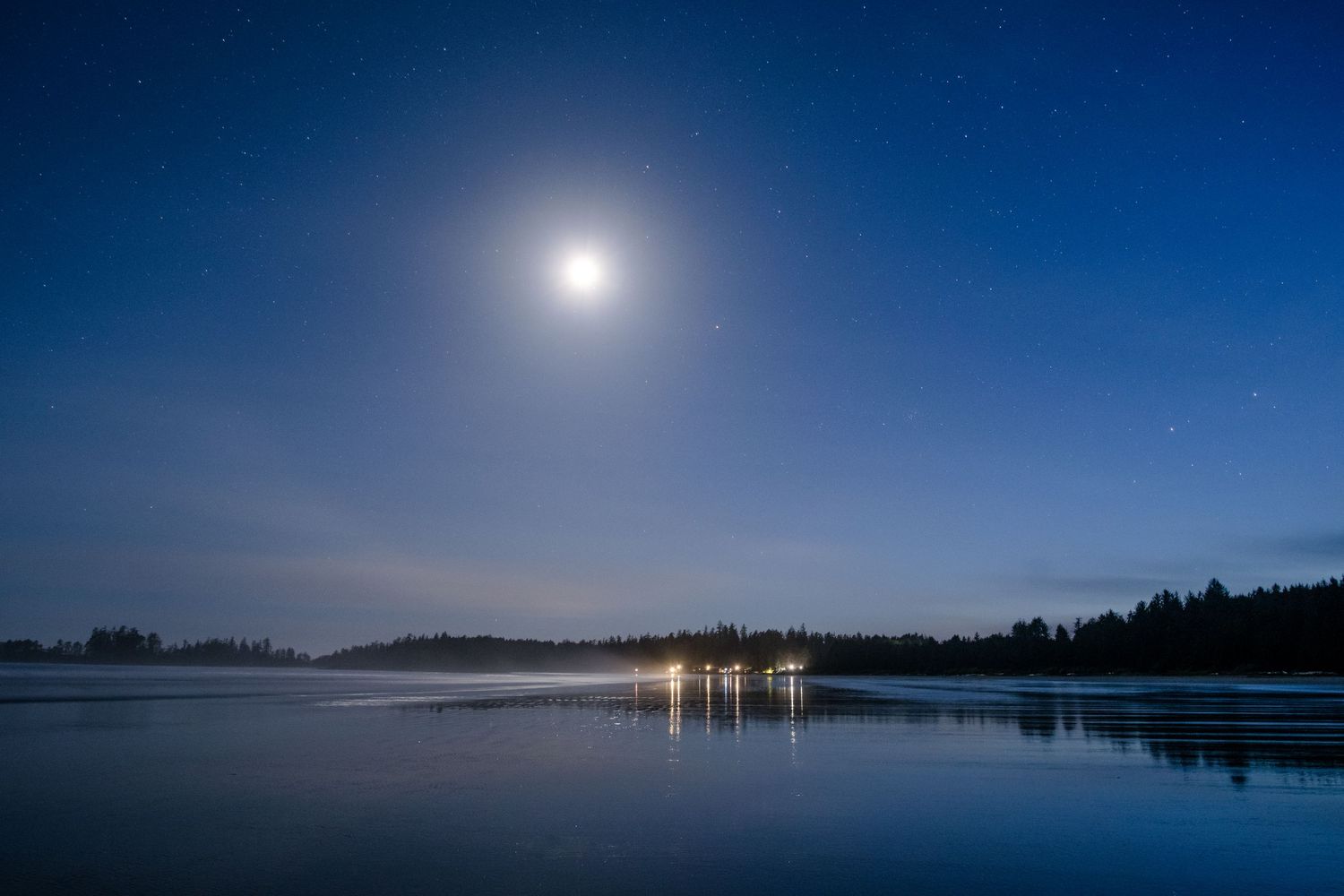 Panoramic nighttime seascape with full moon reflecting on calm waters and silhouetted shoreline.
