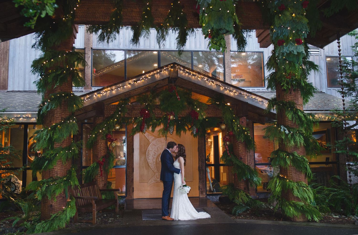 A couple stands in a romantic garden setting with lush greenery and elegant architecture at sunset.