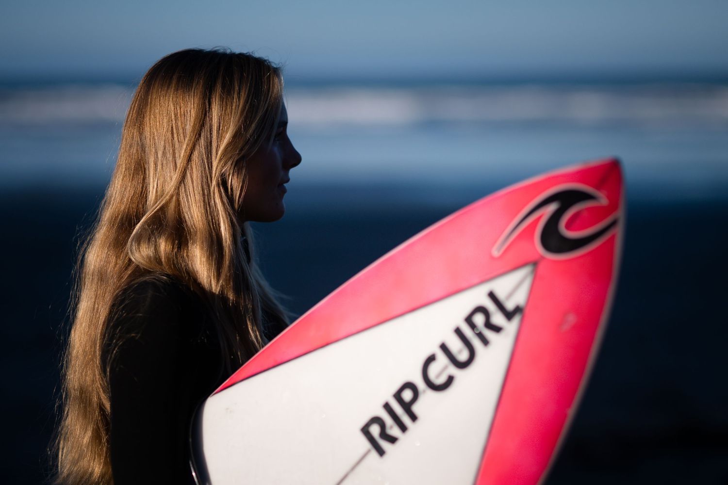 Surfer carrying pink surfboard walks along misty beach at sunset.