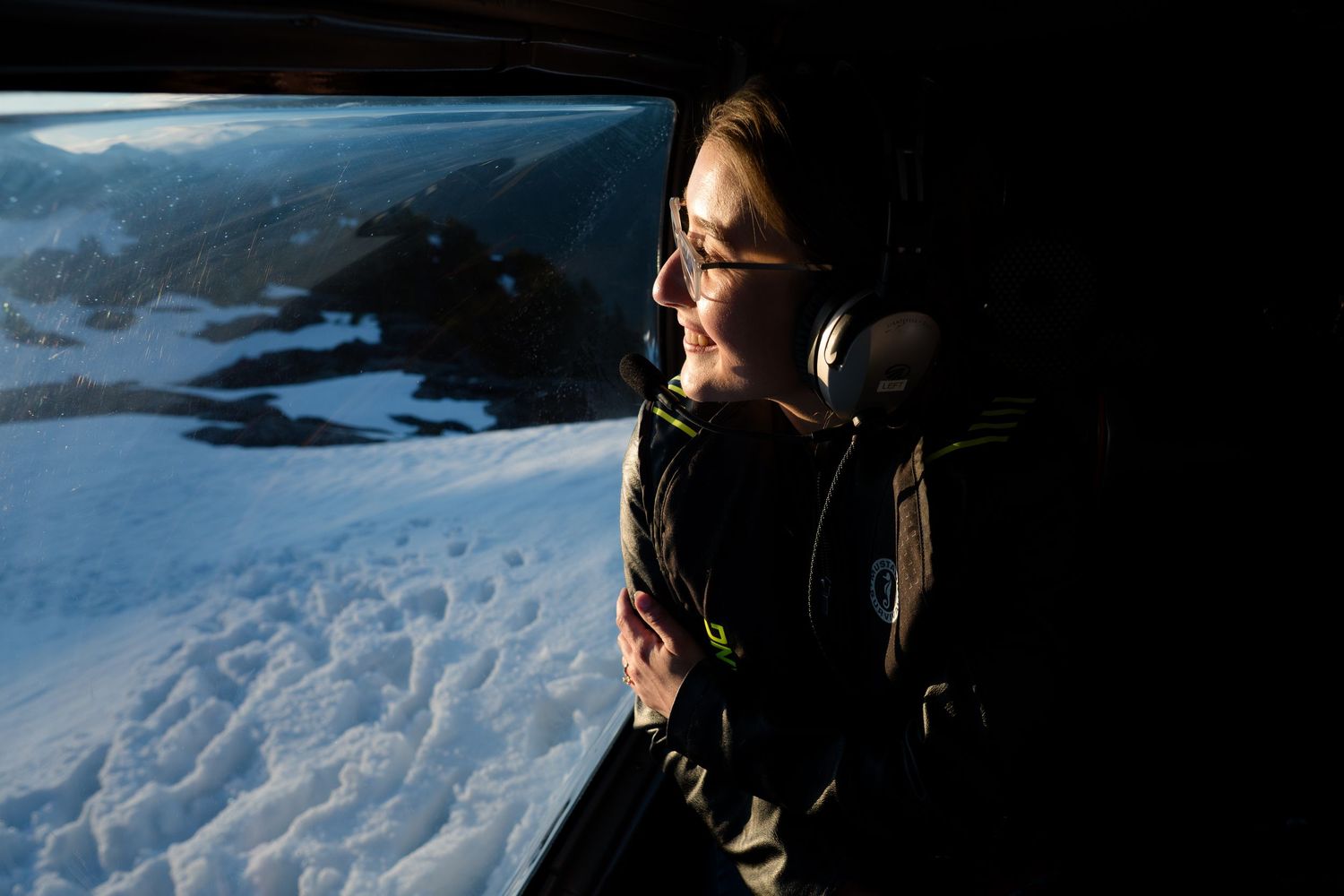 Woman wearing headphones gazing out helicopter window at breathtaking snowy mountain landscape below.