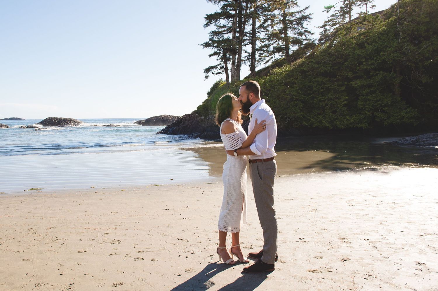 Couple sharing intimate moment on scenic Pacific Northwest beach surrounded by evergreen trees.
