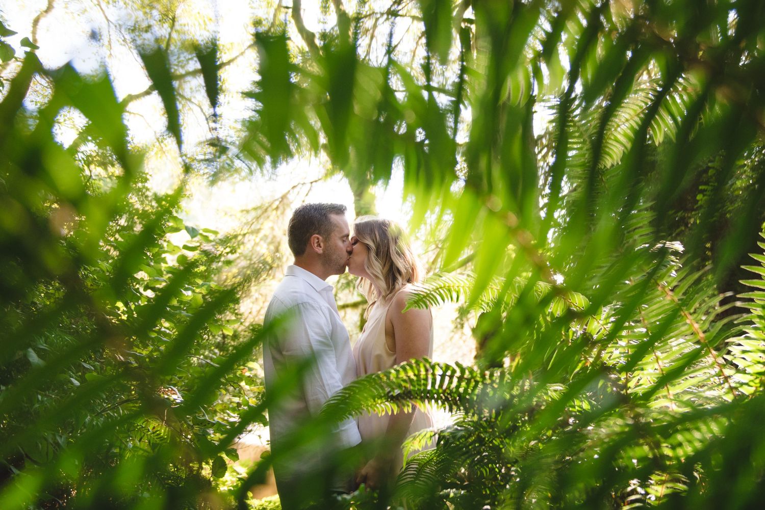 Romantic couple embraces among lush green ferns in a garden setting with soft natural lighting filtering through leaves.