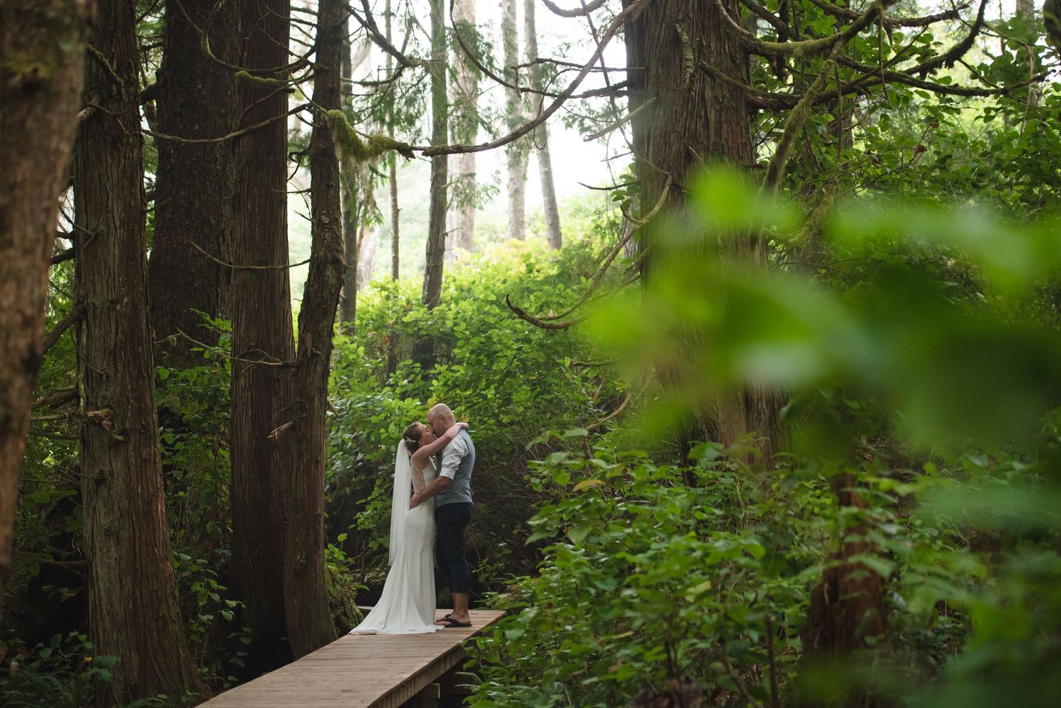 A couple stands together on a wooden boardwalk surrounded by lush green forest.