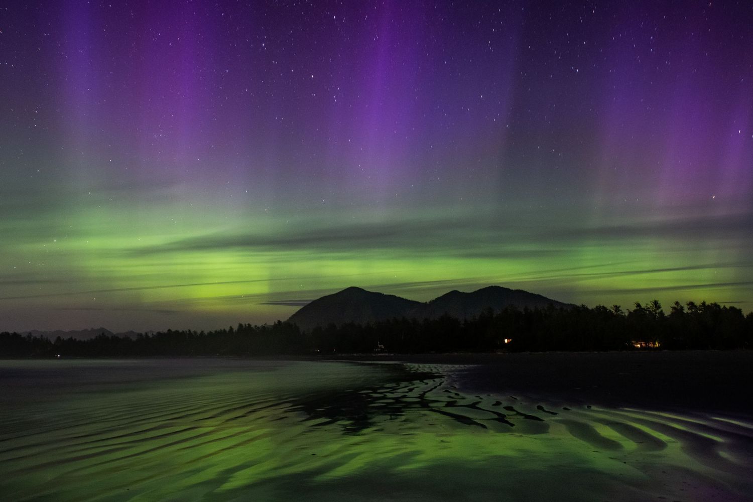 Purple and green aurora borealis lights dance over a dark mountain landscape reflected in water below.
