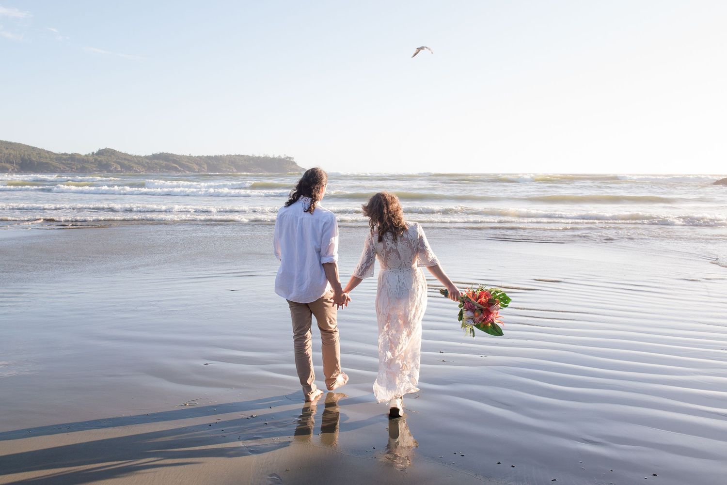 A couple walks along a serene beach at sunset holding hands with ocean waves gently rolling in the background.