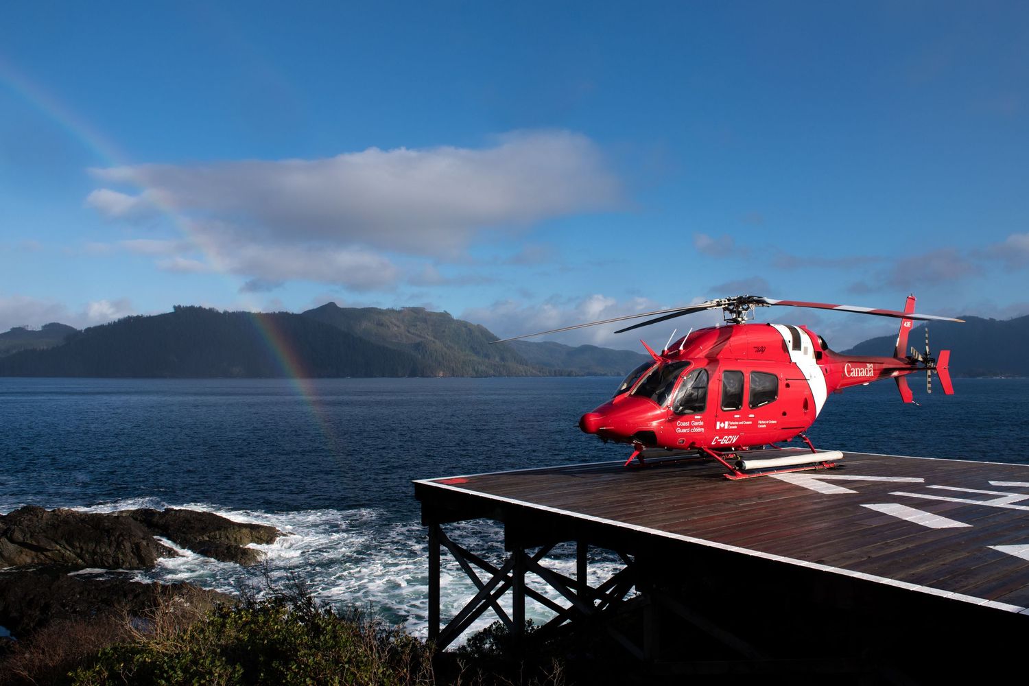 Red rescue helicopter stationed on a coastal helipad with mountains and ocean in the background.