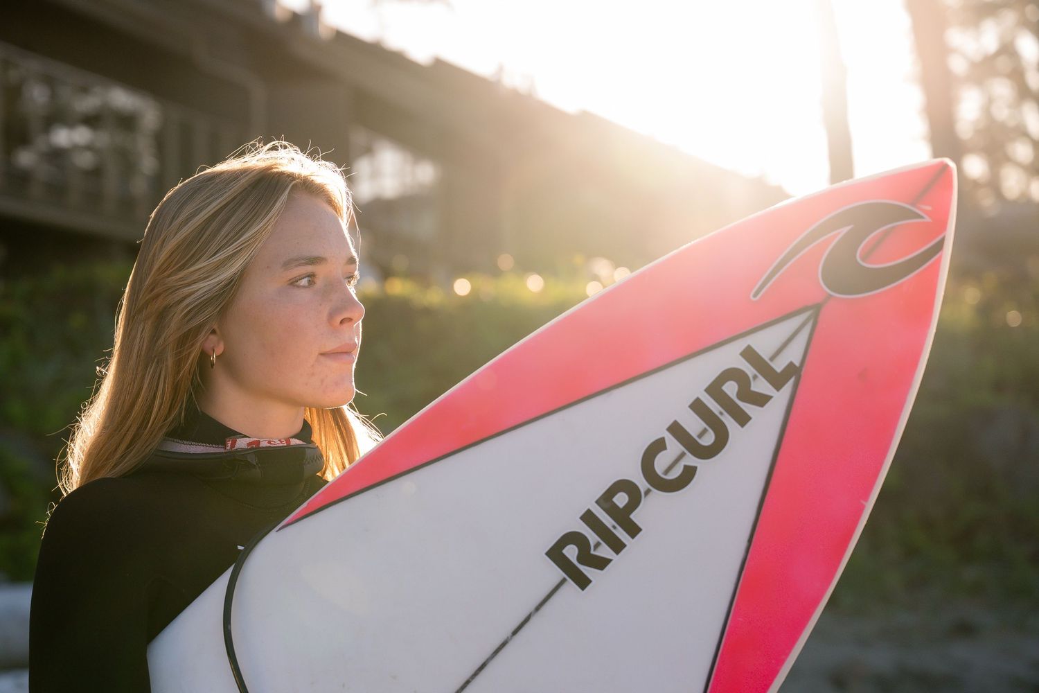A surfer carries a pink and white surfboard along the beach at sunset creating a silhouetted profile.