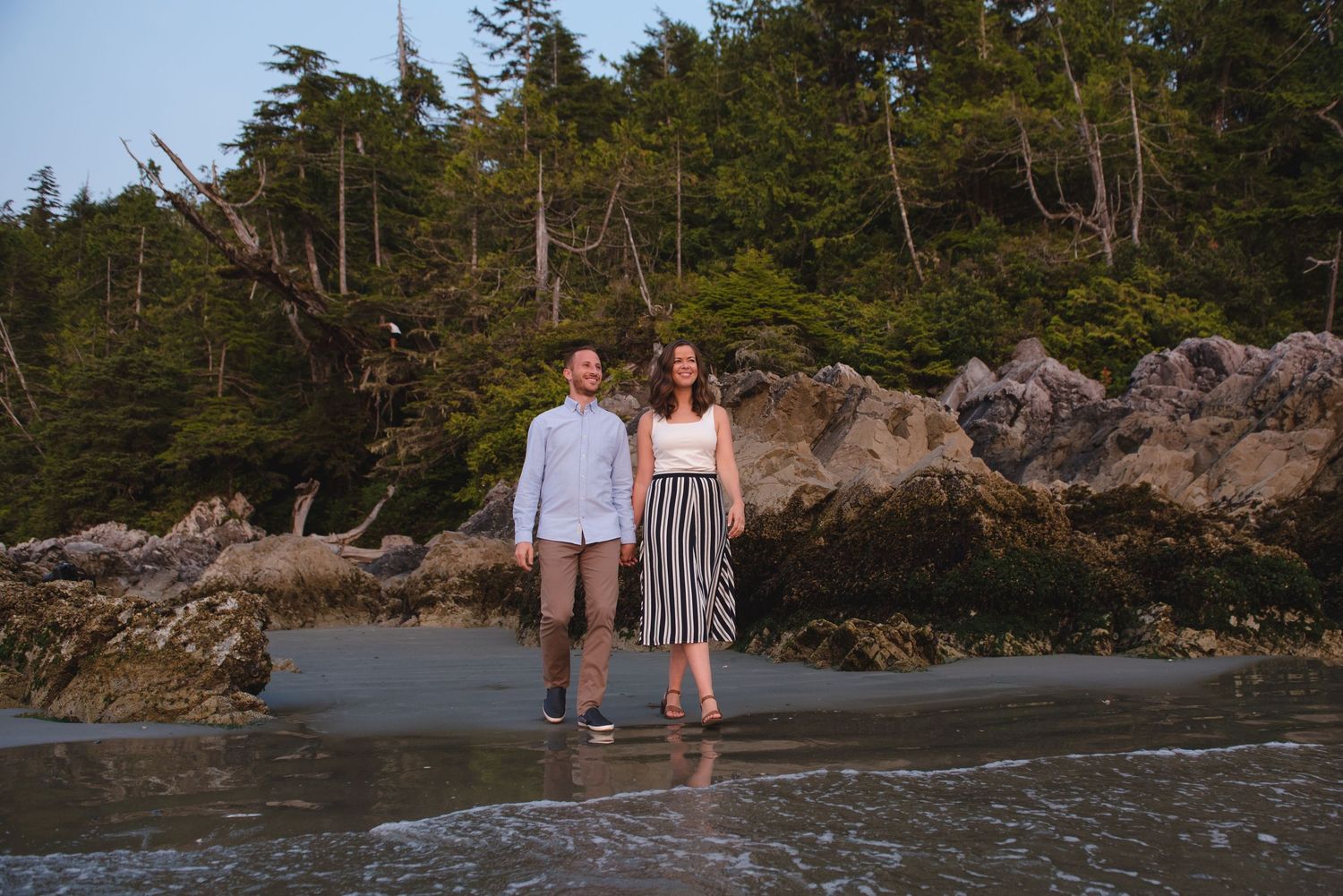 A couple stands together on a rocky beach with evergreen trees in the background.