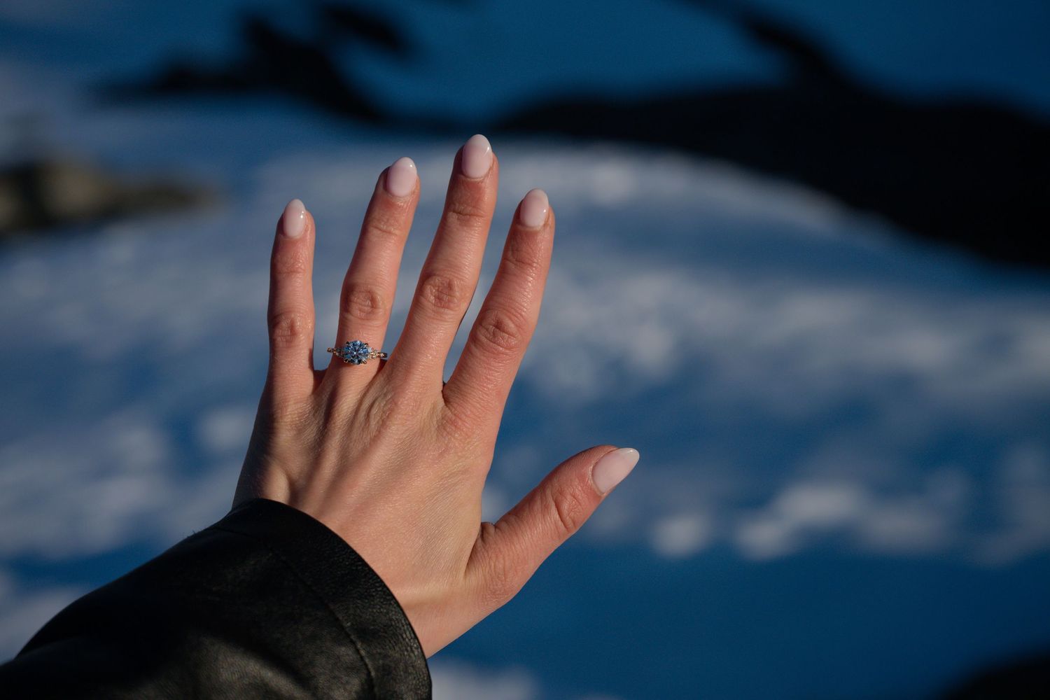 Close-up of a womans hand displaying a beautiful engagement ring with light blue gemstone against a snowy backdrop.