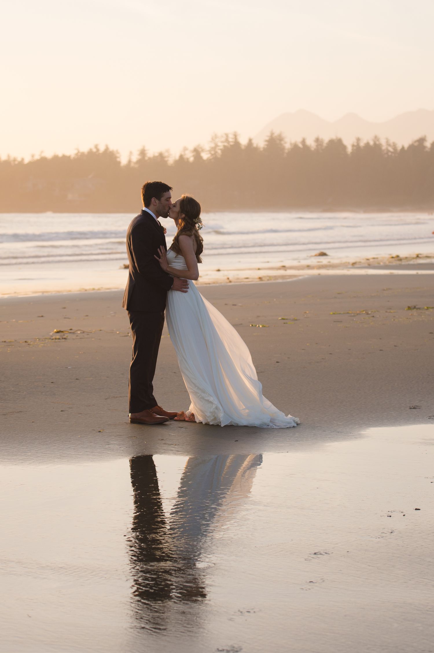 Couple in formal attire dancing on beach at sunset with waves and forest in background.