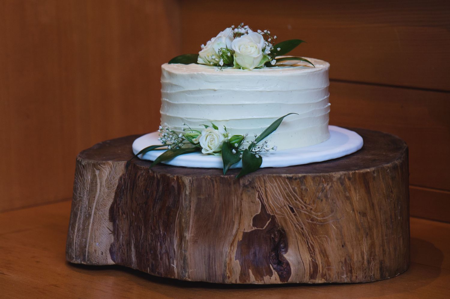 White wedding cake displayed on rustic wood slice platter against brown background.