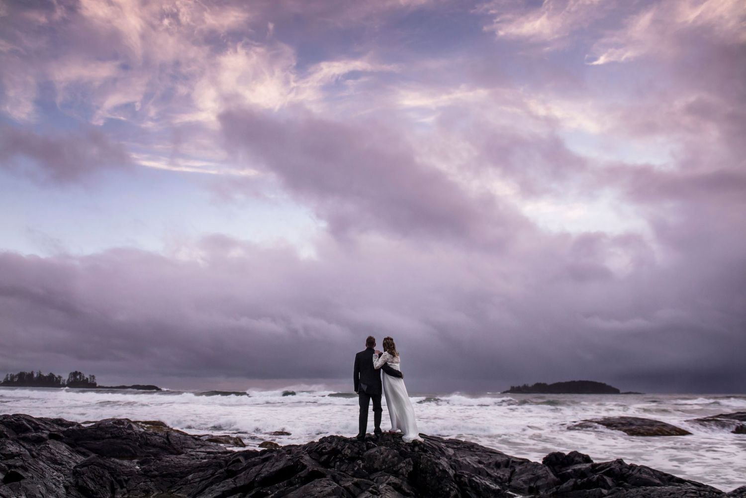 Dramatic cloudy sky over rugged coastal rocks with silhouetted figures watching crashing waves at sunset in Tofino.