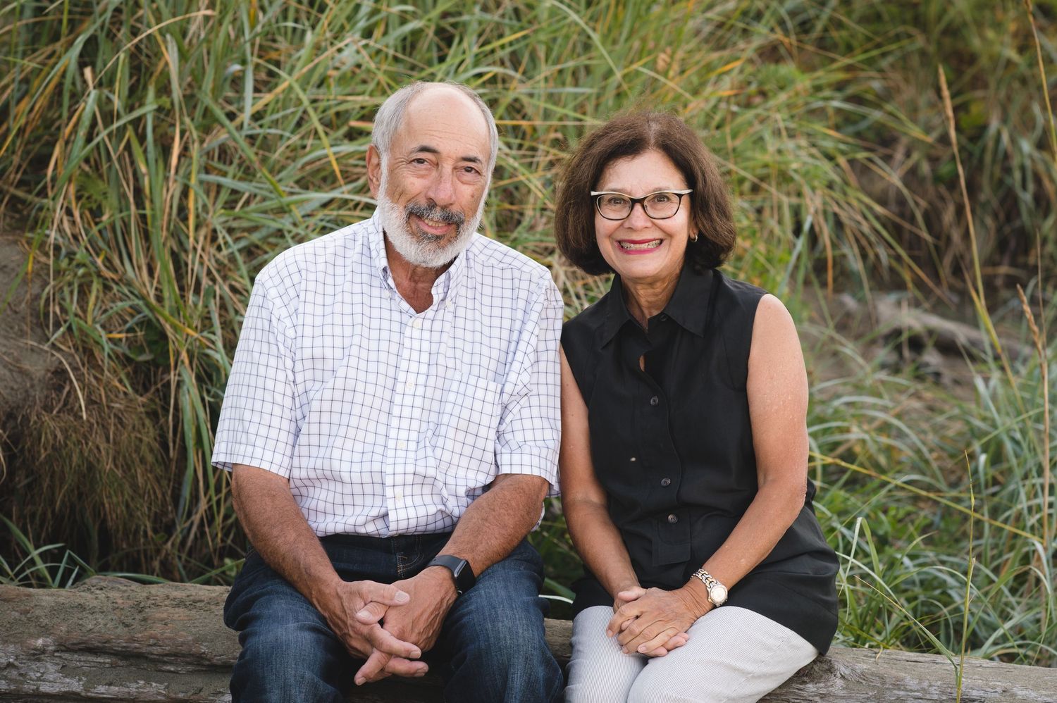 A mature couple sits together on driftwood surrounded by beach grass.