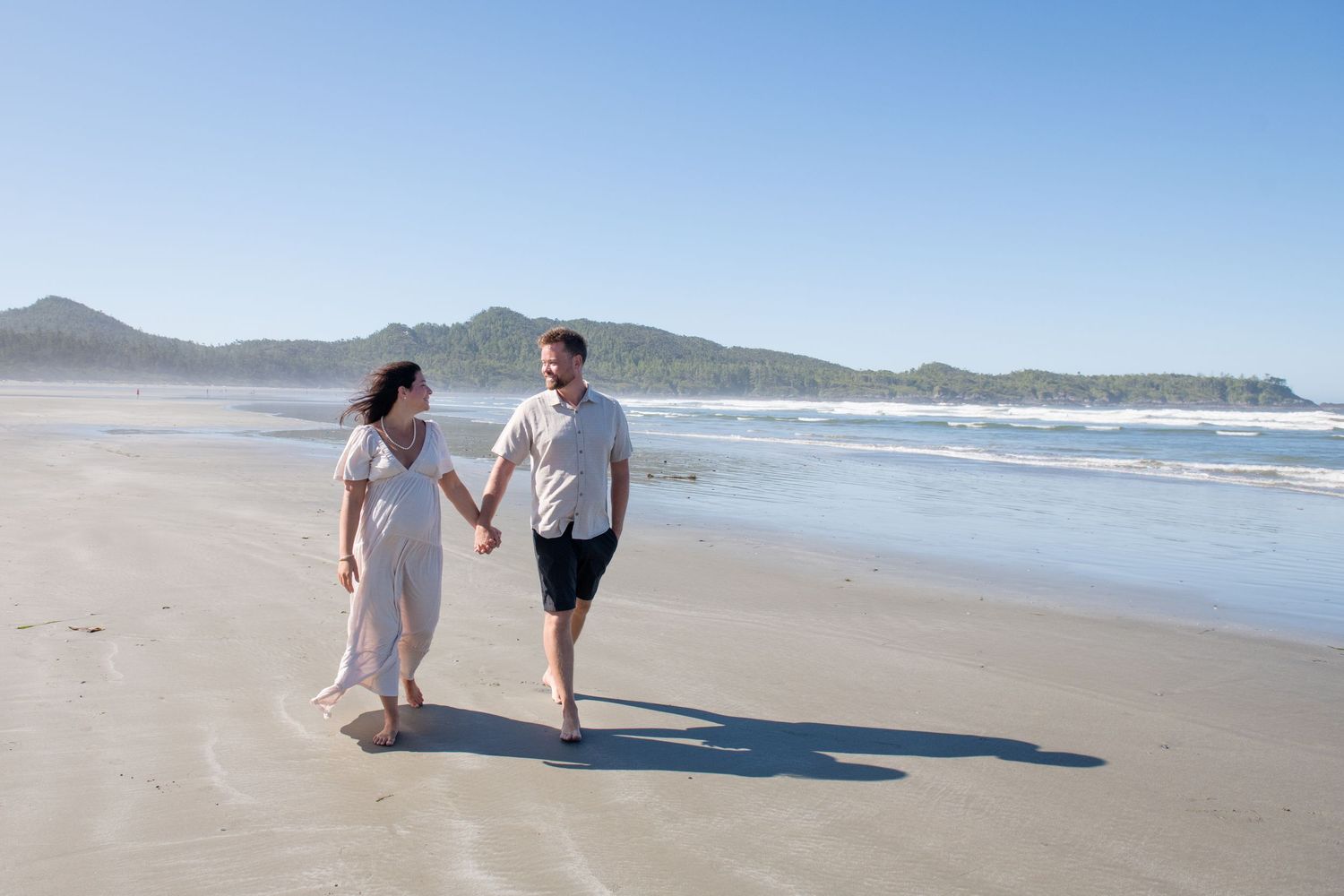 A couple walks hand in hand along a scenic beach with mountains and ocean waves in the background.