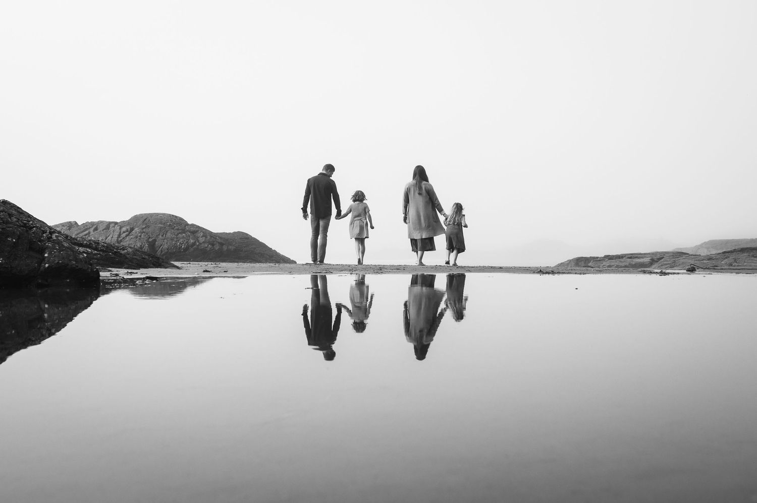 Silhouettes of people walking on wet beach with mountains in distance during foggy day.