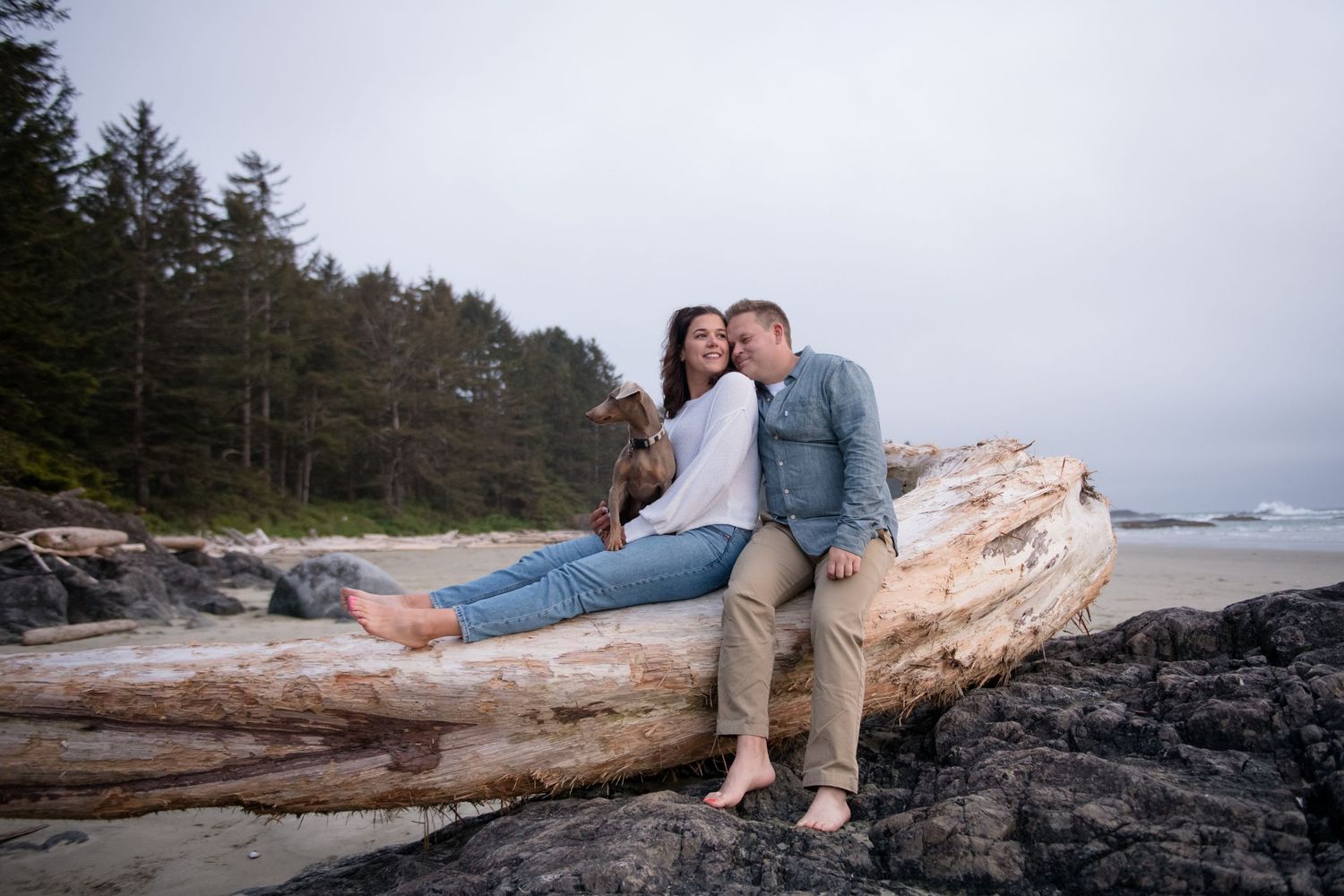 A couple sits together on driftwood at the rocky Pacific Northwest coastline.