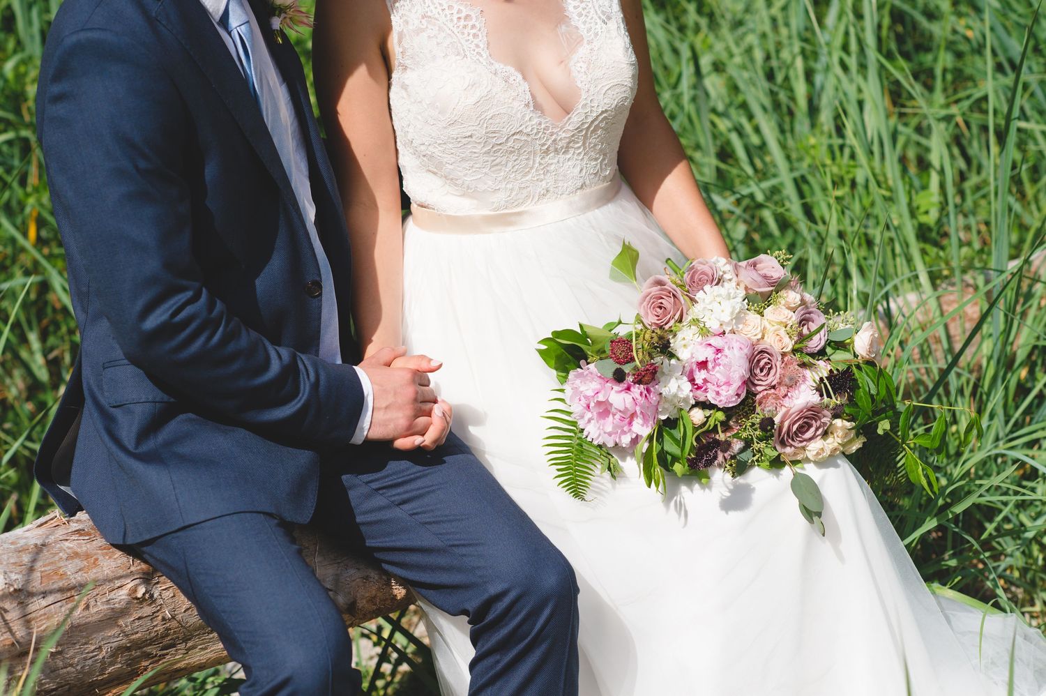 Couple in wedding attire poses together in a lush green field with pink floral bouquet.