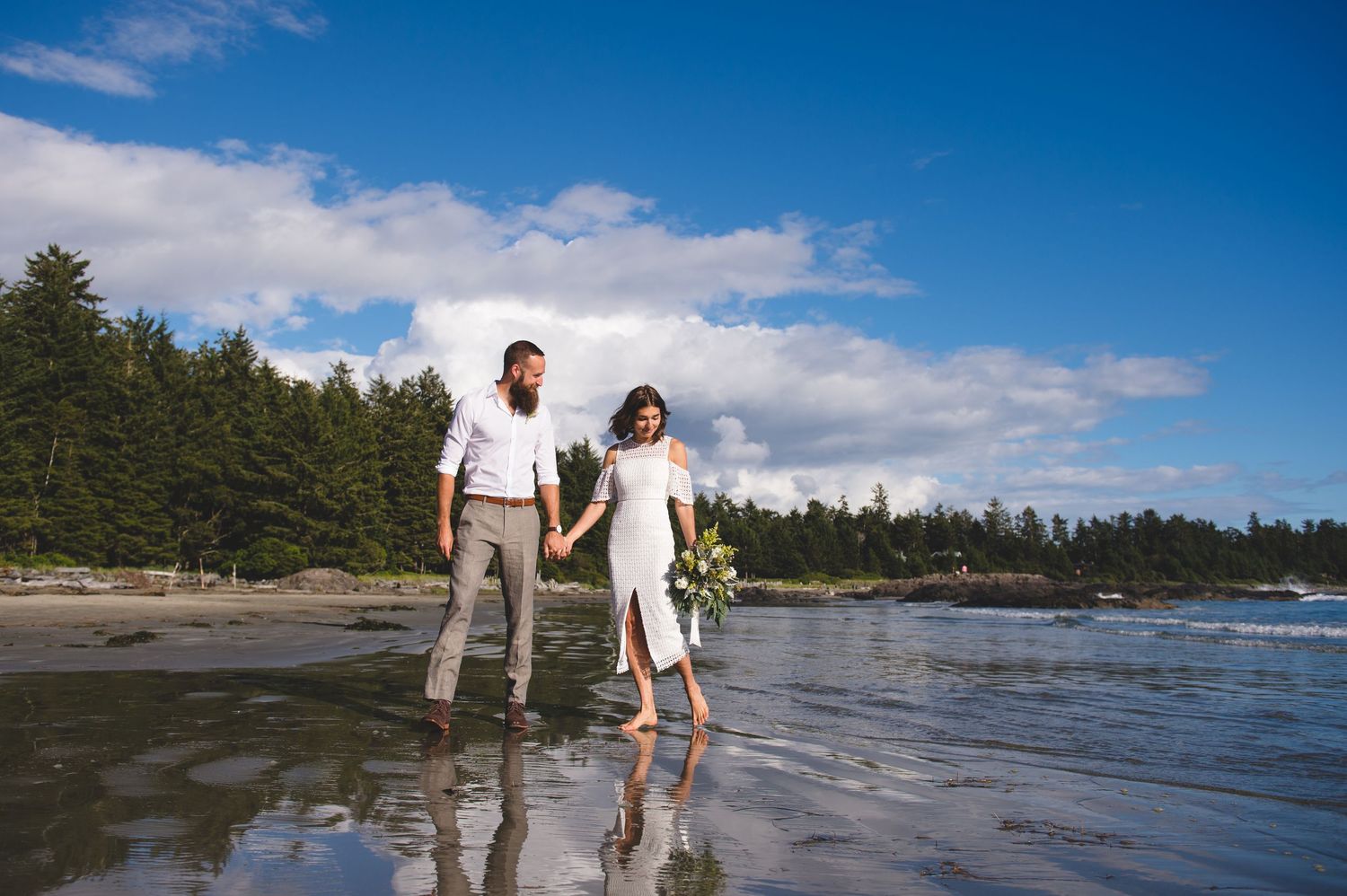 Couple walking along rocky Pacific Northwest coastline with waves crashing against shore on sunny day.