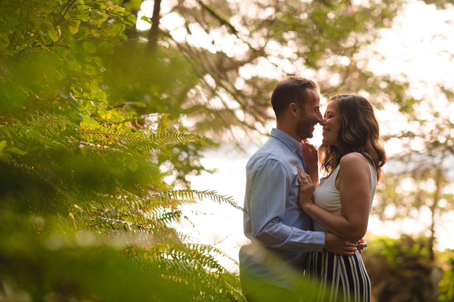 Couple shares intimate moment surrounded by sunlit trees and greenery.