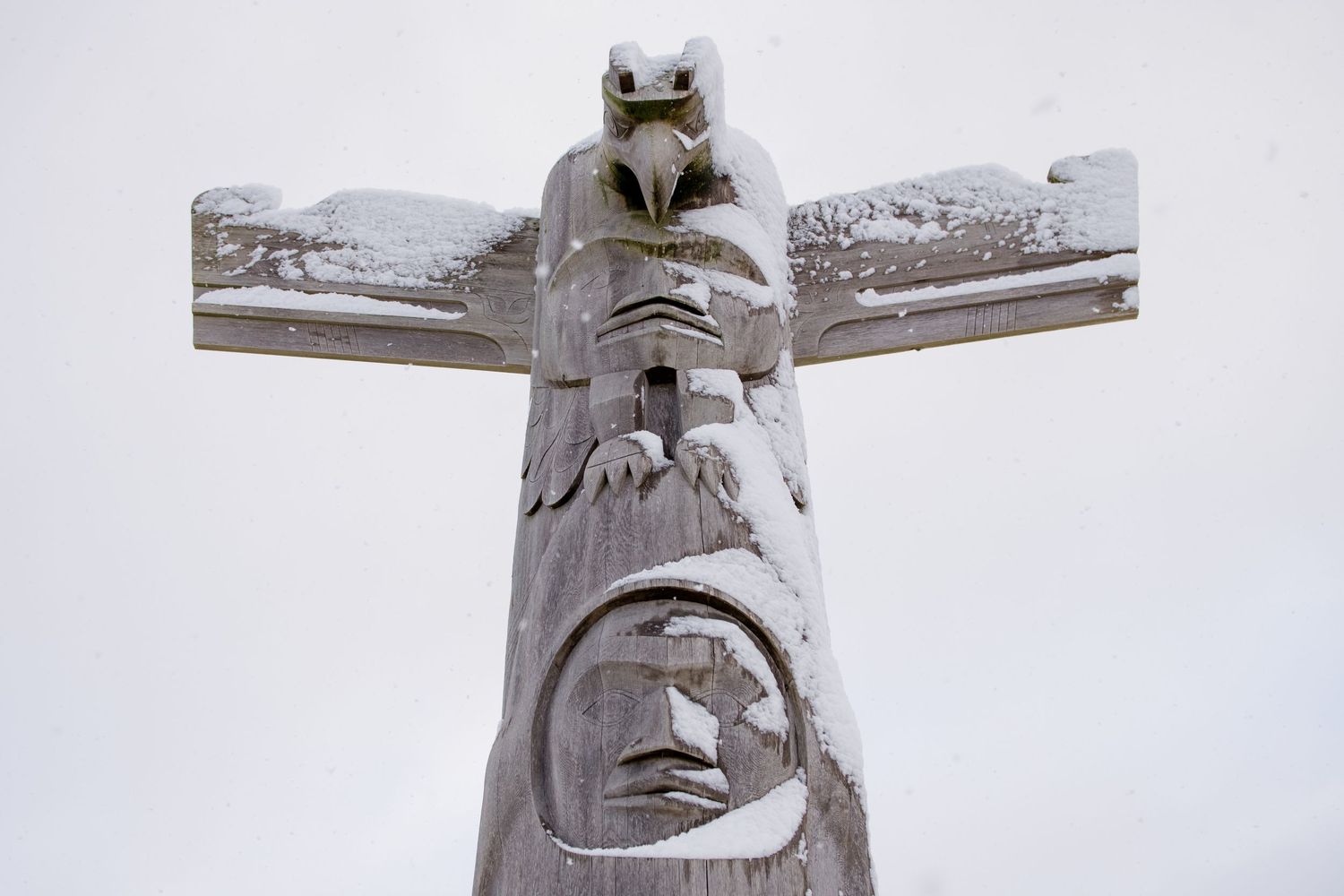 Close-up view of a carved wooden totem pole with faces and traditional Native American designs against a light background.