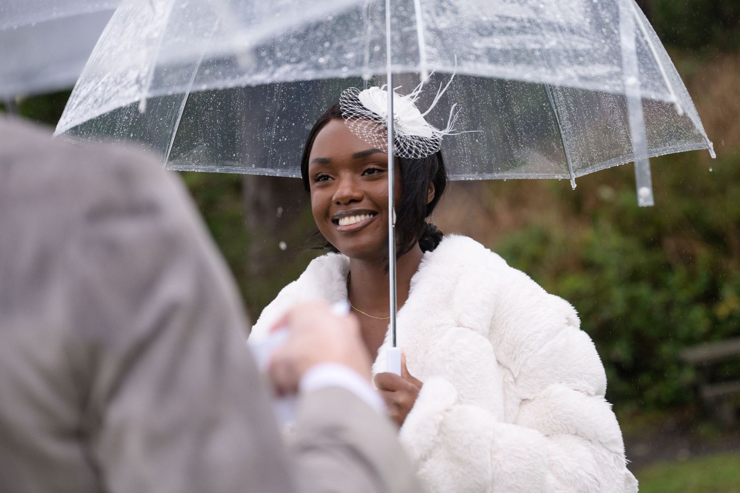 A bride in a white wedding dress holds a clear umbrella while standing outdoors in the rain.